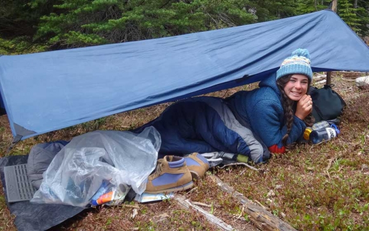 a person smiles from under a tarp shelter amongst trees on an outward bound course